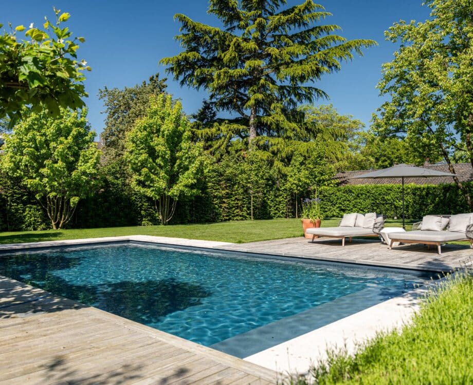 Moderne tuin met zwembad in Nijmegen. Strakke moderne tuin met groot gazon. Vlonderterras met ligbedjes, parasol. Grote bomen voor natuurlijke schaduw. Strak modern zwembad. Heerlijk zwemmen in de zomer. Tuinontwerp en tuinaanleg door Uw Tuin hoveniers uit Malden.