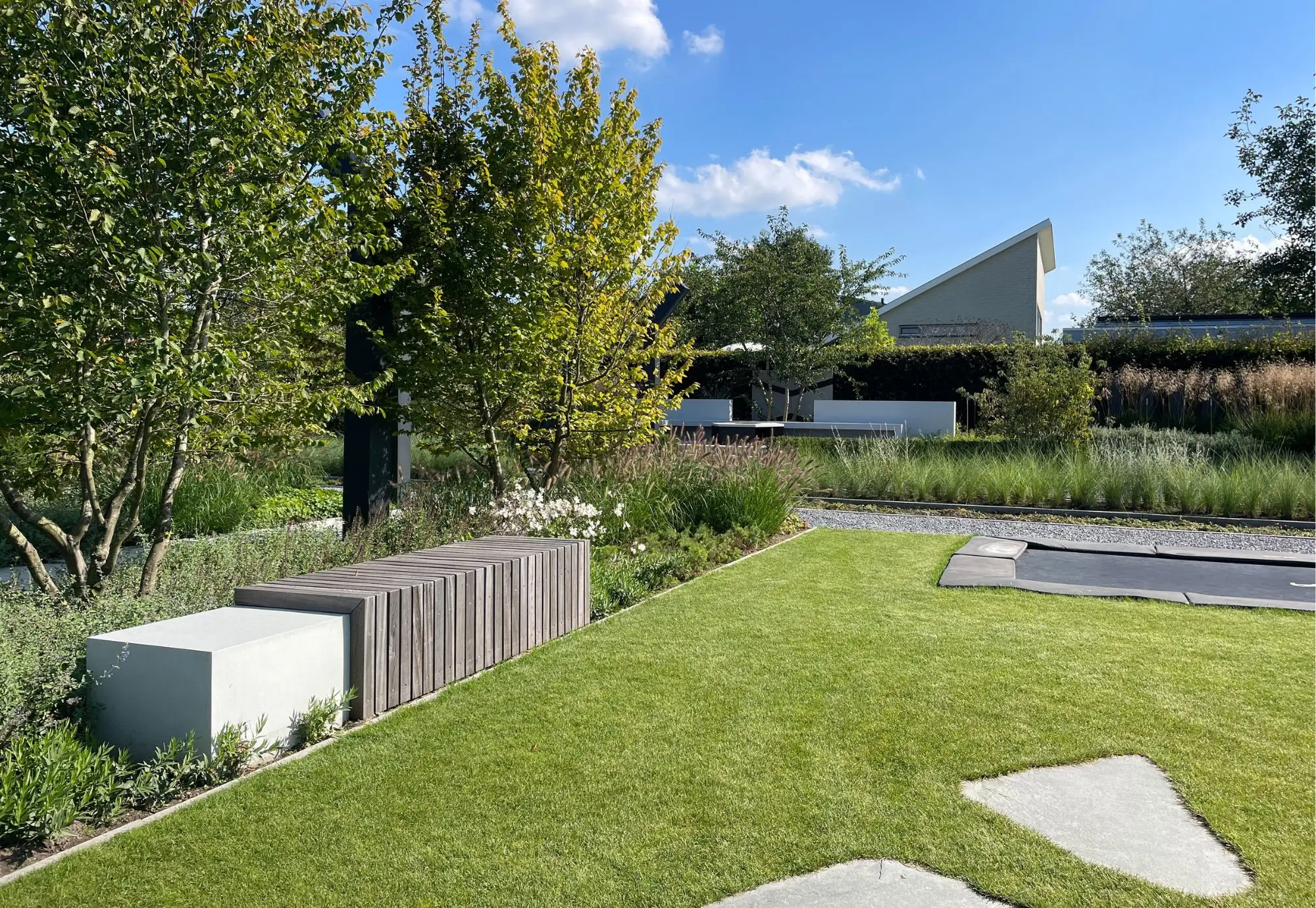 Designtuin in Beuningen met zwembad. Groot gazon met stapstenen. Trampoline voor de kinderen in het groen. Zitelement wit met houten bekleding in plantvak. Plantvak met strakke randen, weelderige beplanting, natuurlijk uiterlijk. Solitaire heesters geven karakter aan de tuin. Tuinontwerp en Tuinaanleg door Uw Tuin hoveniers uit Malden.