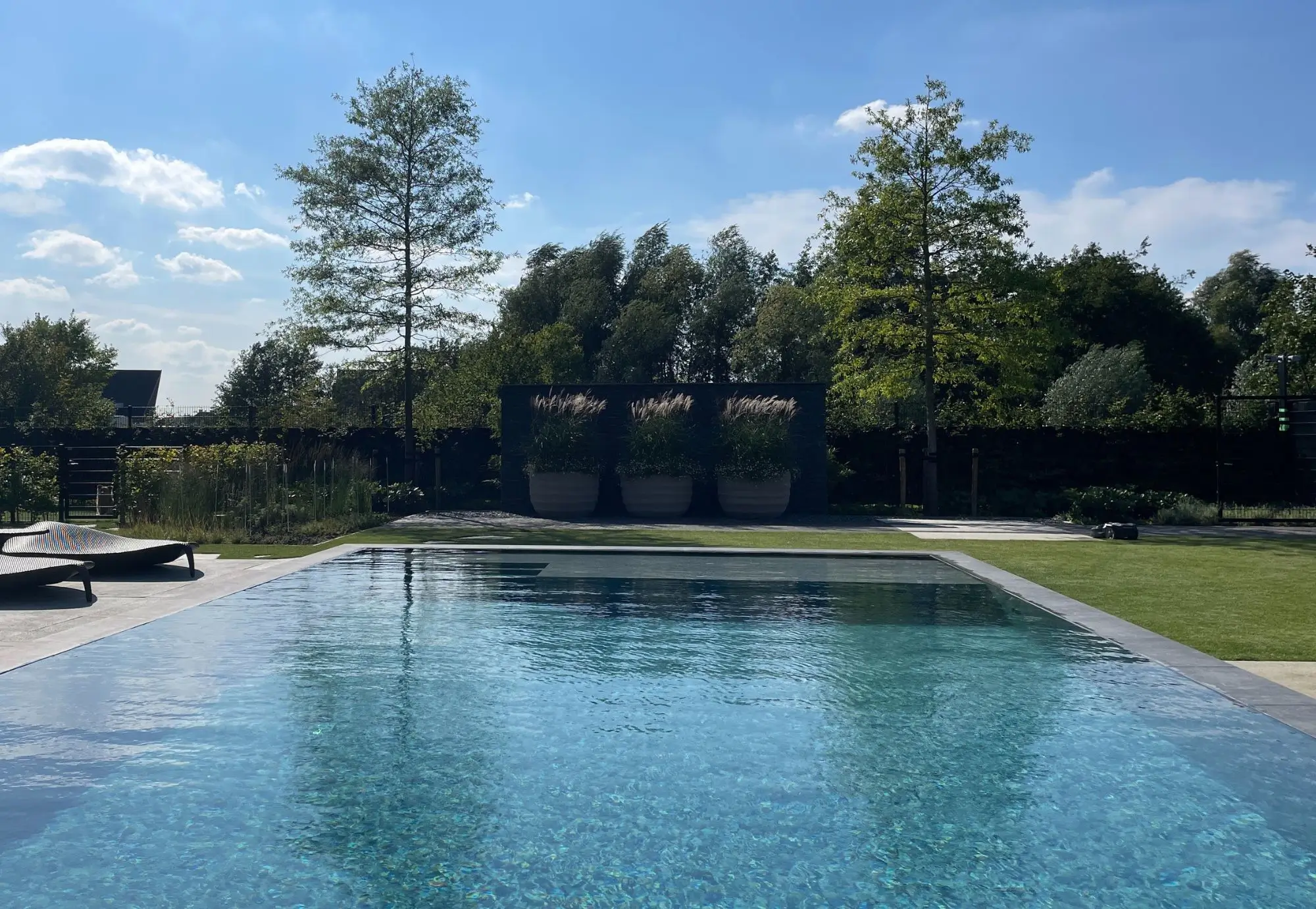 Vooraanzicht van een modern zwembad met groot vlonderterras met loungeset. Bijzondere Designtuin met een architectonische wand waarvoor drie designpotten staan met beplanting. Groot strak gazon met strakke plantenborders en weelderige beplanting. Solitaire bomen en heesters zorgen voor karakter.