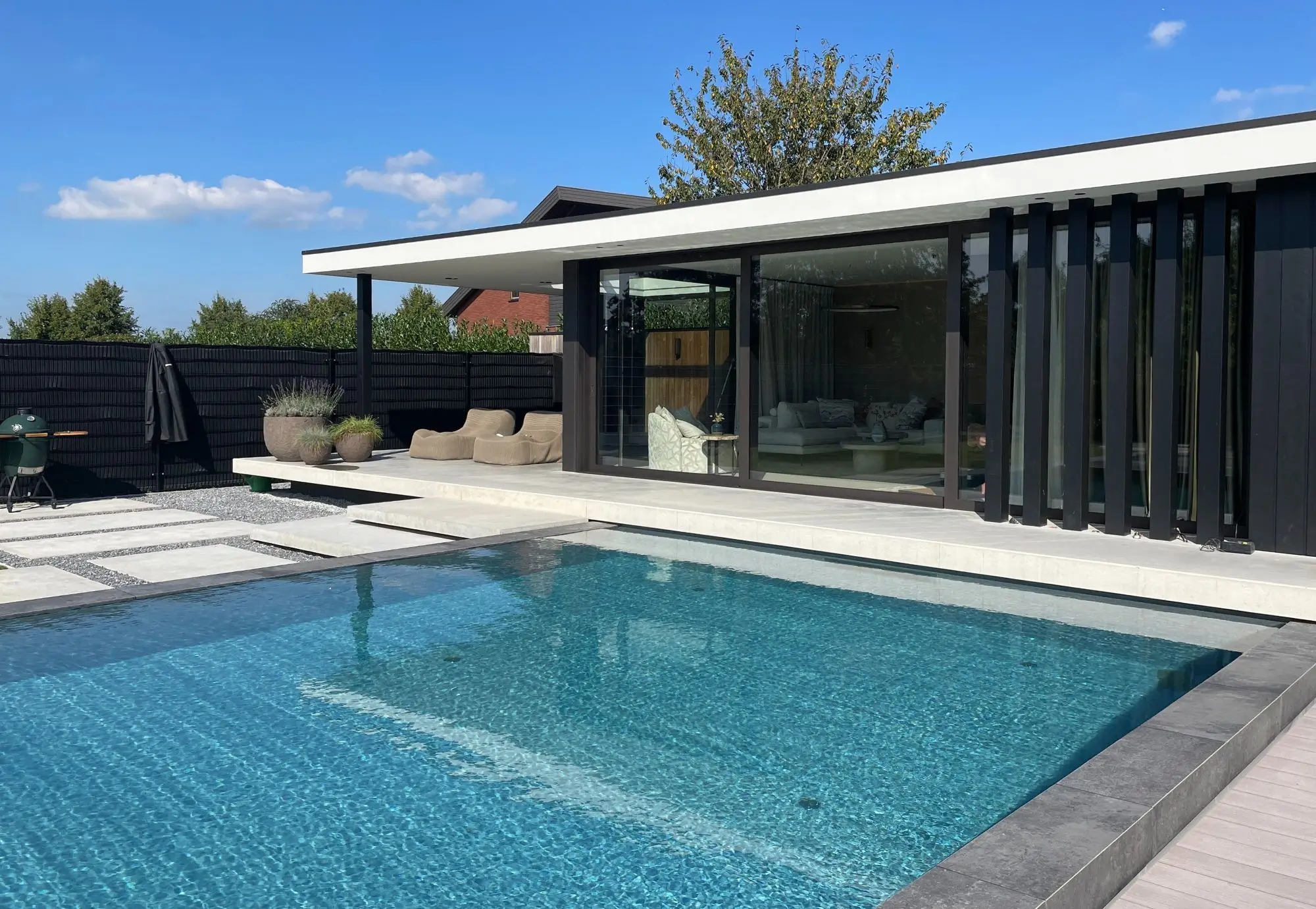 Vooraanzicht van een modern zwembad met modern Pool house, overkapping, tuinhuis. tuinkamer, strakke vormgeving met zwarte Douglas palenwand. Groot keramisch terras met loungeset. Bijzondere Designtuin Stapstenen als trap in het grind, hoogteverschil. Solitaire bomen en heesters zorgen voor karakter. Tuinontwerp en Tuinaanleg door Uw Tuin hoveniers uit Malden.