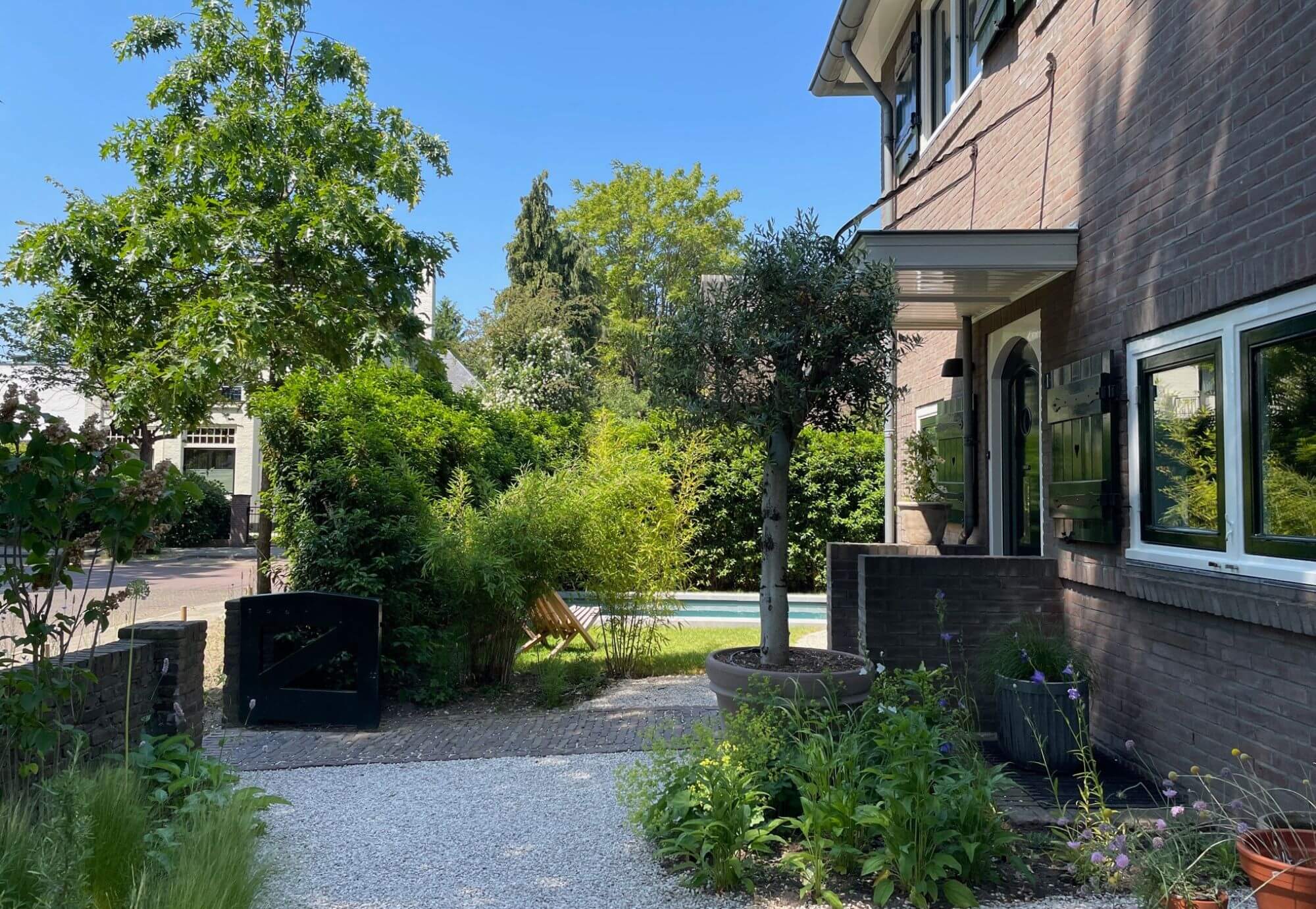 Zijaanzicht van een Stadstuin in Nijmegen met zwembad. Tuinontwerp en tuinaanleg door Uw Tuin hoveniers uit Malden. Organische vormen, klinkerverharding, bestrating, Grind met cortenstalen kantopsluitng. Veel groen. Bestaande hagen, zwembad in groot gazon, jaren 30 woning.