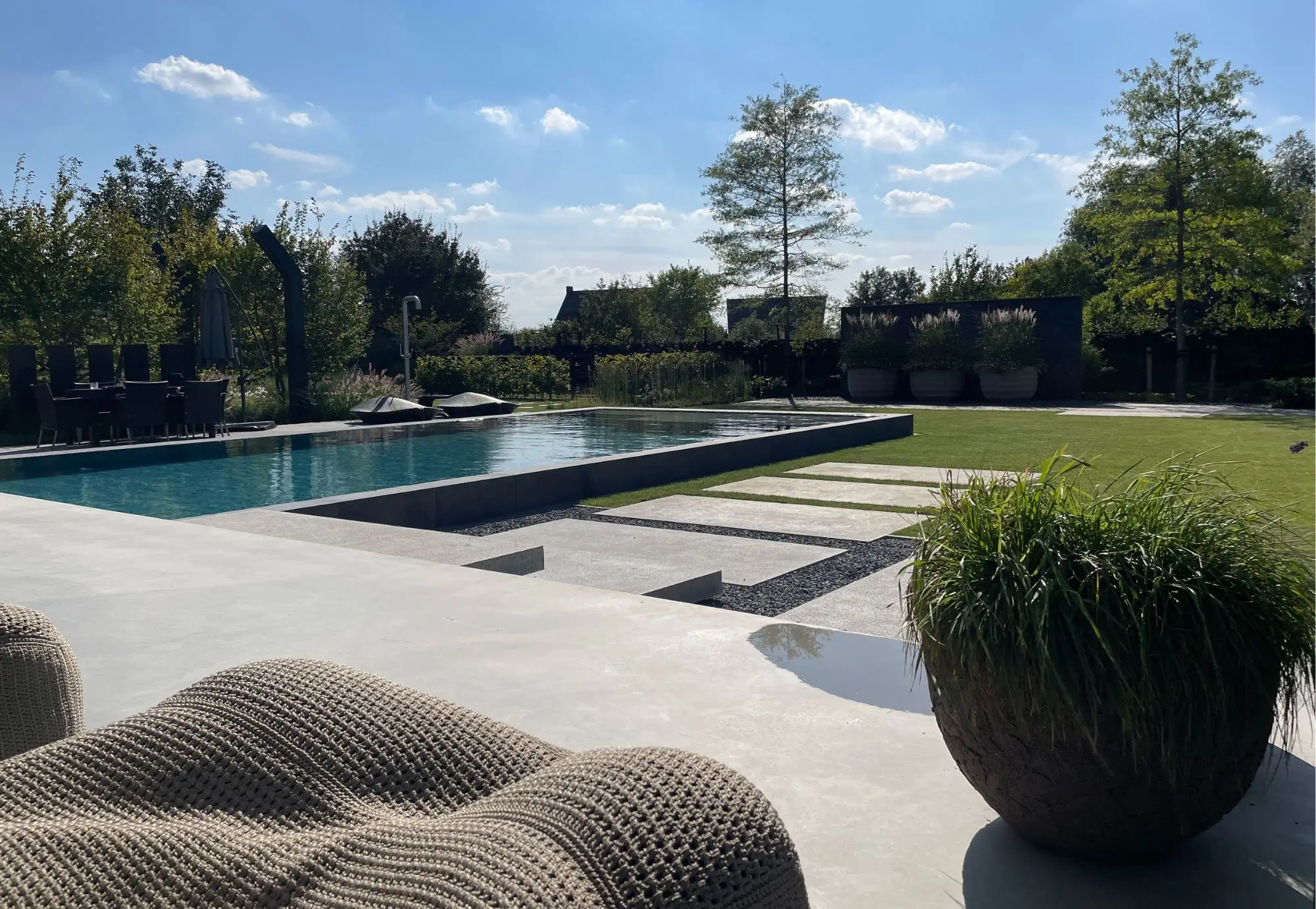 Achtertuin van een Designtuin met modern zwembad. Designtuin met stapstenen in het grind, buitendouche terras en groene borders, groen gazon. Tuinaanleg en Tuinontwerp door Uw Tuin hoveniers uit Malden.