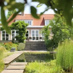 Doorkijk naar woonhuis i Mook. Natuurlijke zwemvijver met strakke vorm, groene oevers, Vlonderpad en staptegels als springplank. Tuinontwerp en tuinaanleg door Uw Tuin hoveniers uit Malden.